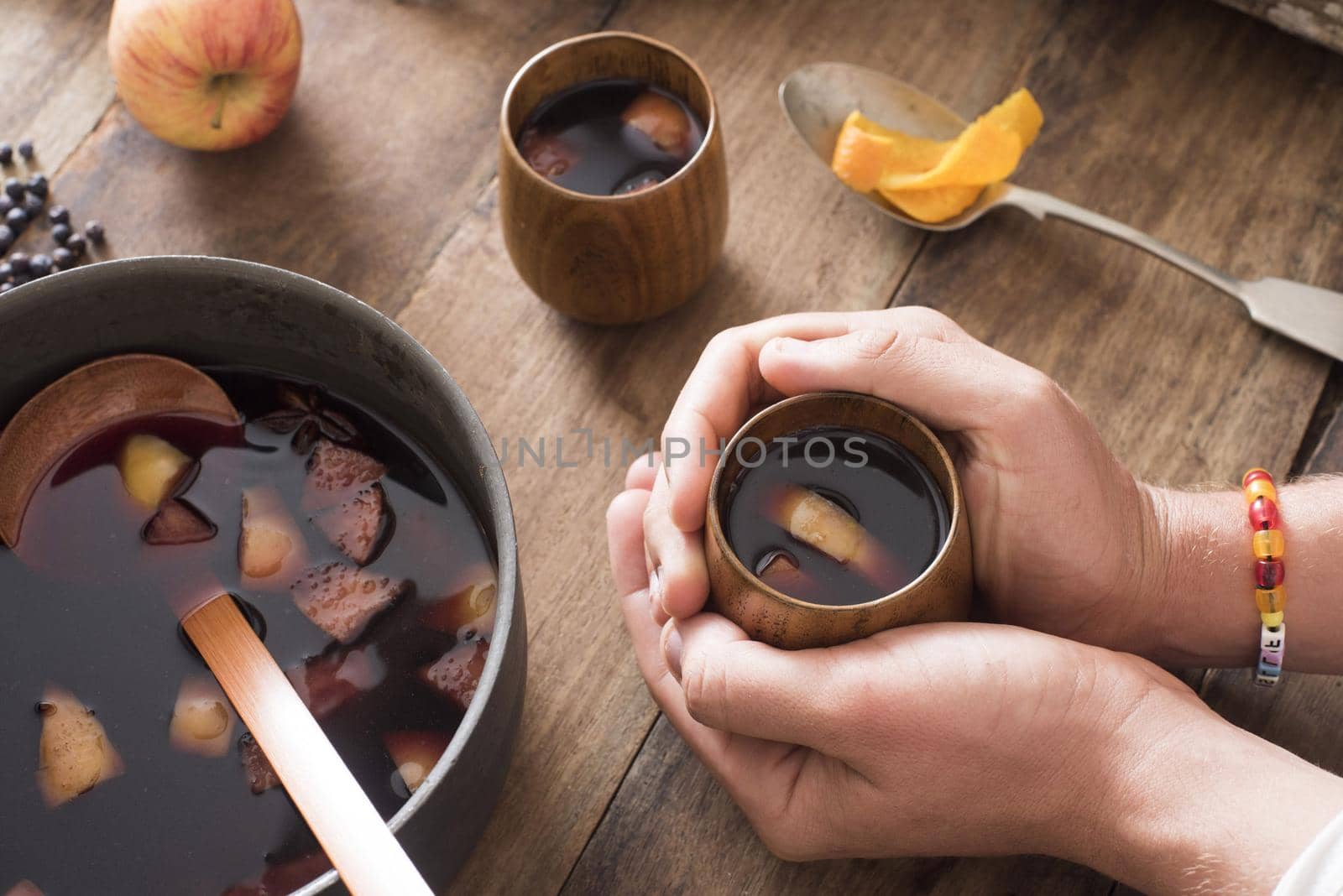 Person warming their hands on a mug of spicy fruity mulled wine seated at a table with a pot of freshly made brew