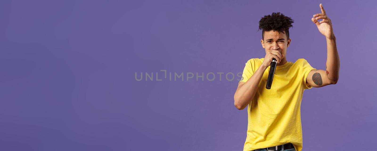 Portrait of young carefree sassy hipster guy with tattoos, yellow t-shirt performing in front audience with his hip-hop or rap, singing song, raising finger up, hold microphone, karaoke by Benzoix