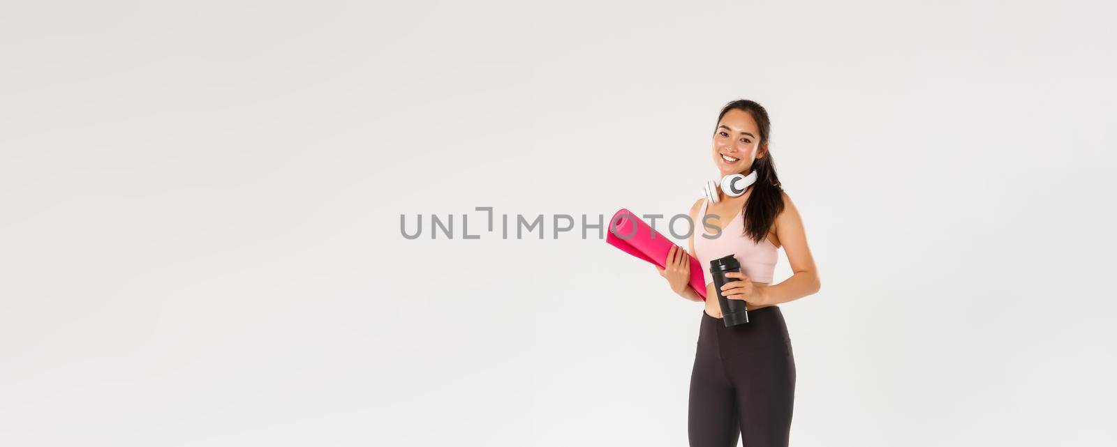 Full length of smiling slim and beautiful asian female athlete, girl carry fitness exercise rubber mat and water bottle, going for yoga classes or workout in gym with own equipment, white background by Benzoix