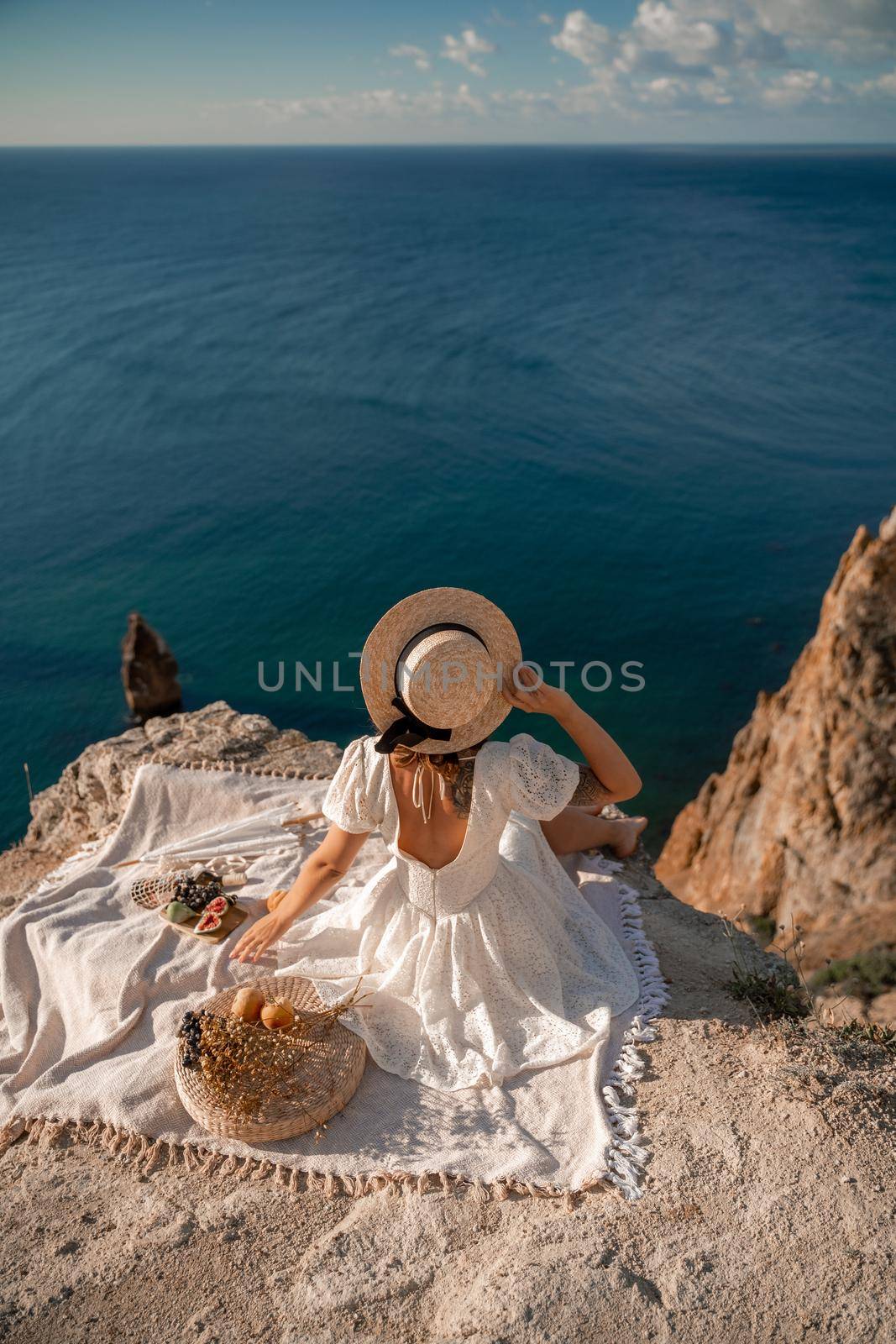 Street photo of a beautiful woman with dark hair in a white dress and hat having a picnic on a hill overlooking the sea.