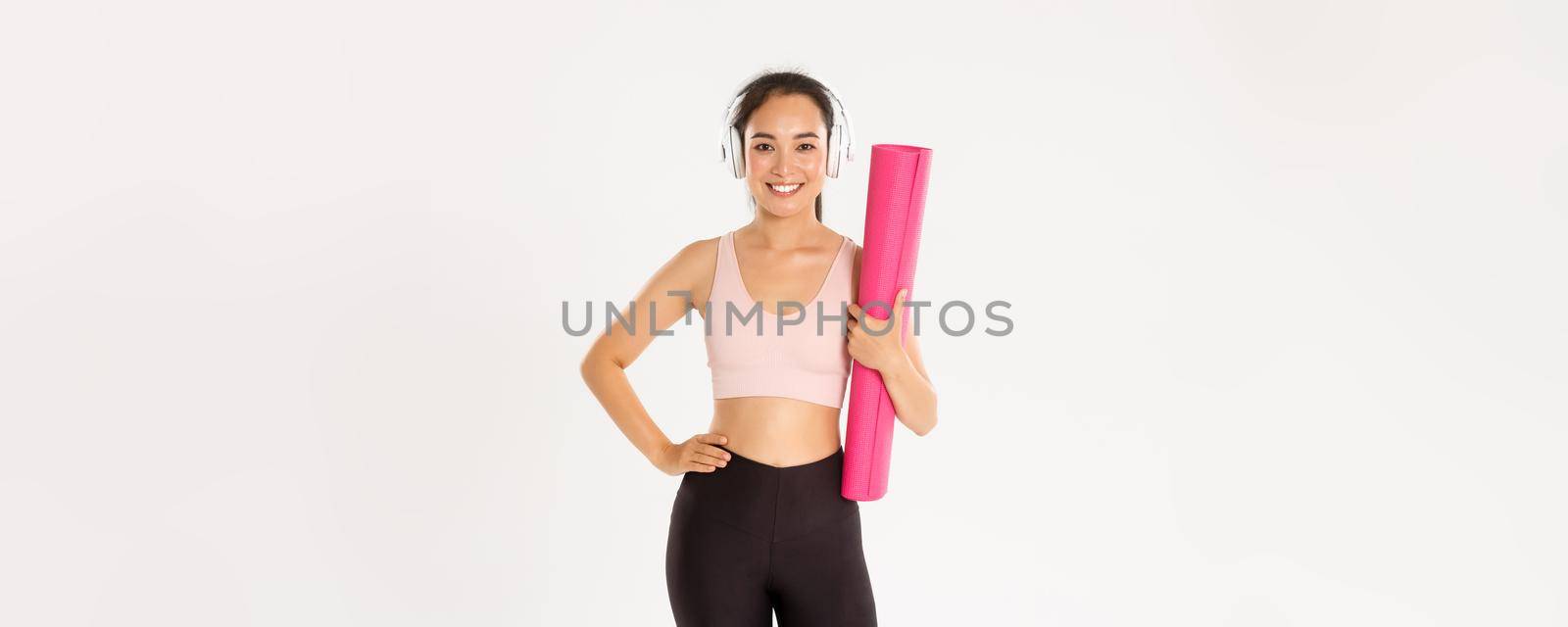 Sport, wellbeing and active lifestyle concept. Sassy beautiful asian fitness girl, woman ready for yoga classes or home training, holding rubber mat for workout and listening music in headphones by Benzoix