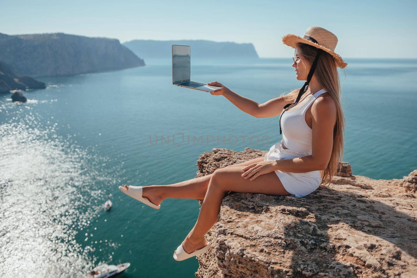 Successful business woman in yellow hat working on laptop by the sea. Pretty lady typing on computer at summer day outdoors. Freelance, travel and holidays concept.