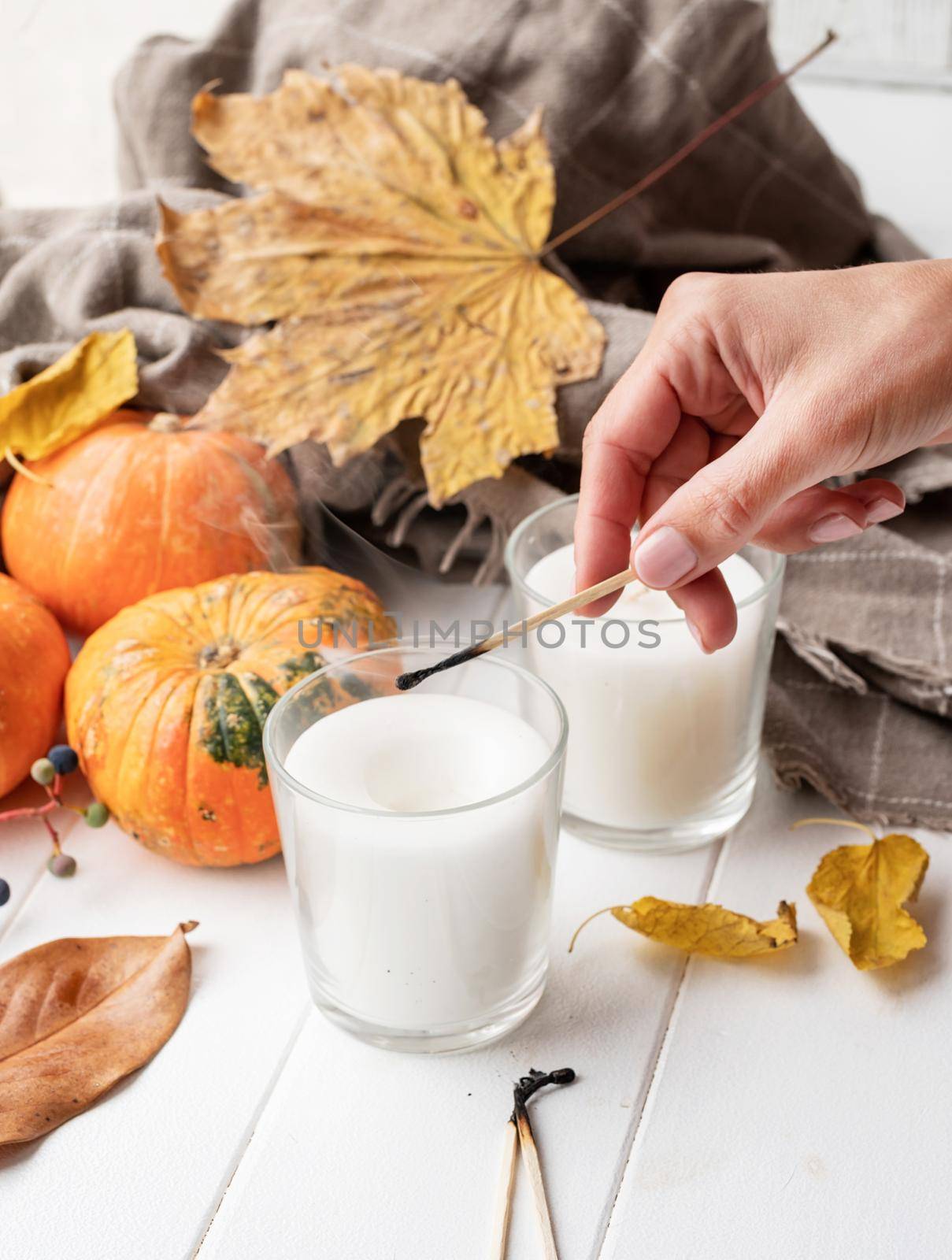 Hello fall. Cozy warm image. High angle view autumn composition with sweaters and candles, white candle mockup design. Woman hand lighting candles