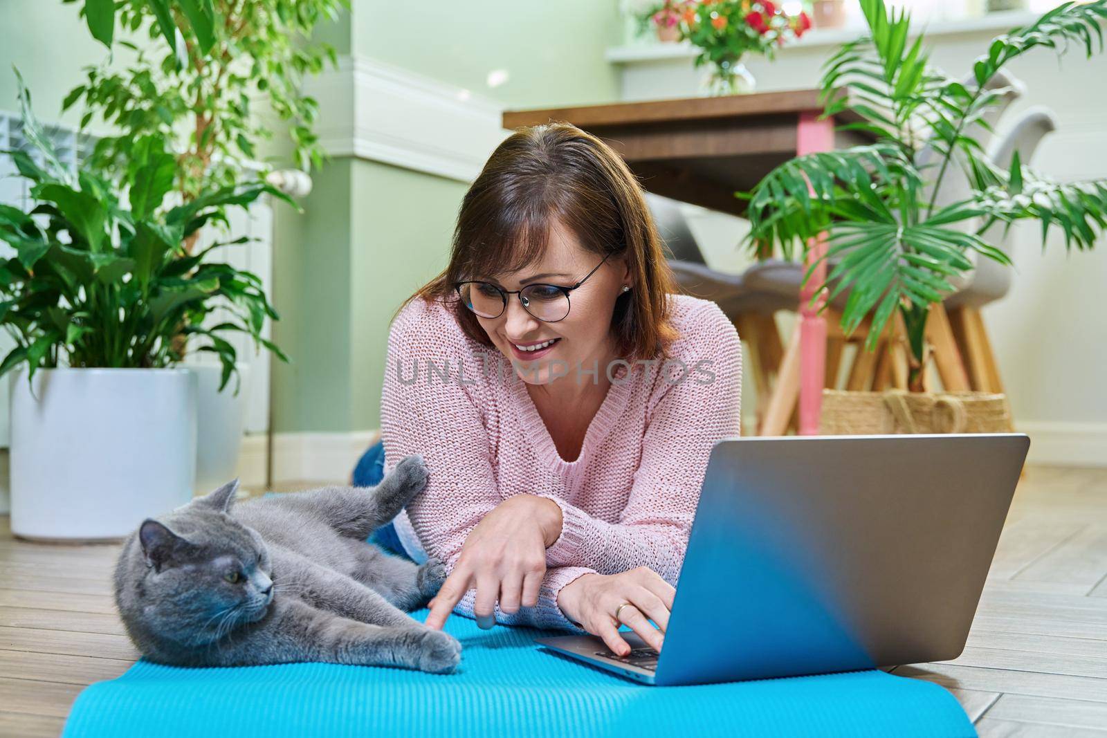 Middle-aged woman lying with cat on exercise mat, looking at laptop by VH-studio