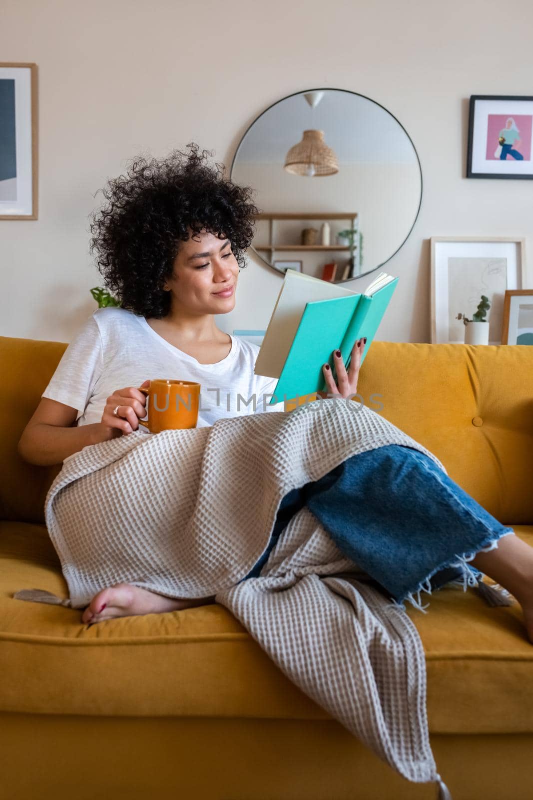 Relaxed, happy African american woman at home living room sitting on the couch, reading a book and drinking tea. Lifestyle concept.