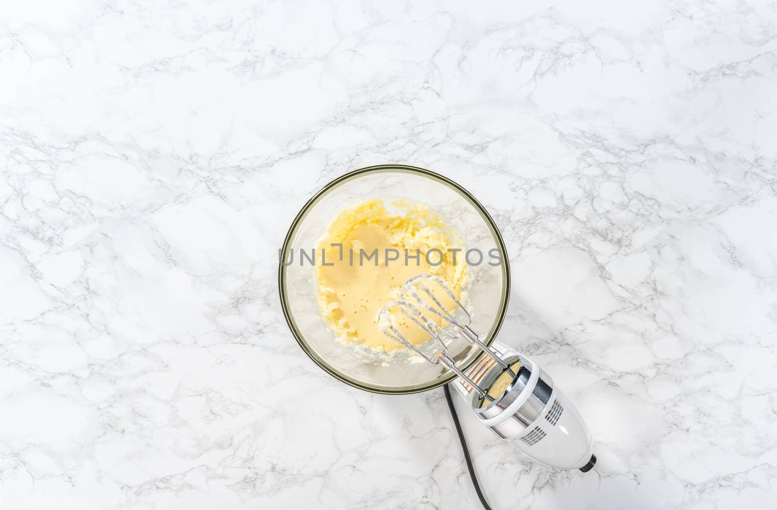 Flat lay. Mixing ingredients with a hand mixer in a large mixing bowl to bake mini vanilla cupcakes with ombre pink buttercream frosting.