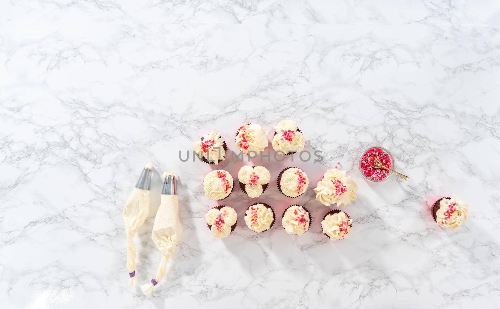 Flat lay. Piping white chocolate ganache frosting on top of red velvet cupcakes and topping with sprinkles.
