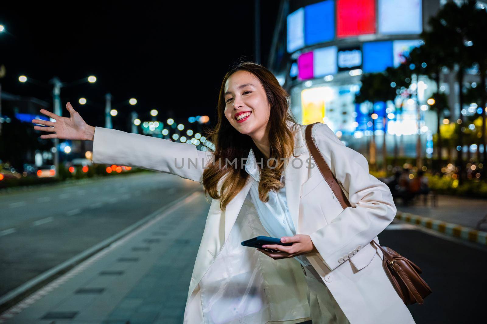 Asian business woman walking to hail waving hand taxi on road in city street at night, Beautiful woman smiling using smartphone application hailing with hand up calling cab outdoor after late work