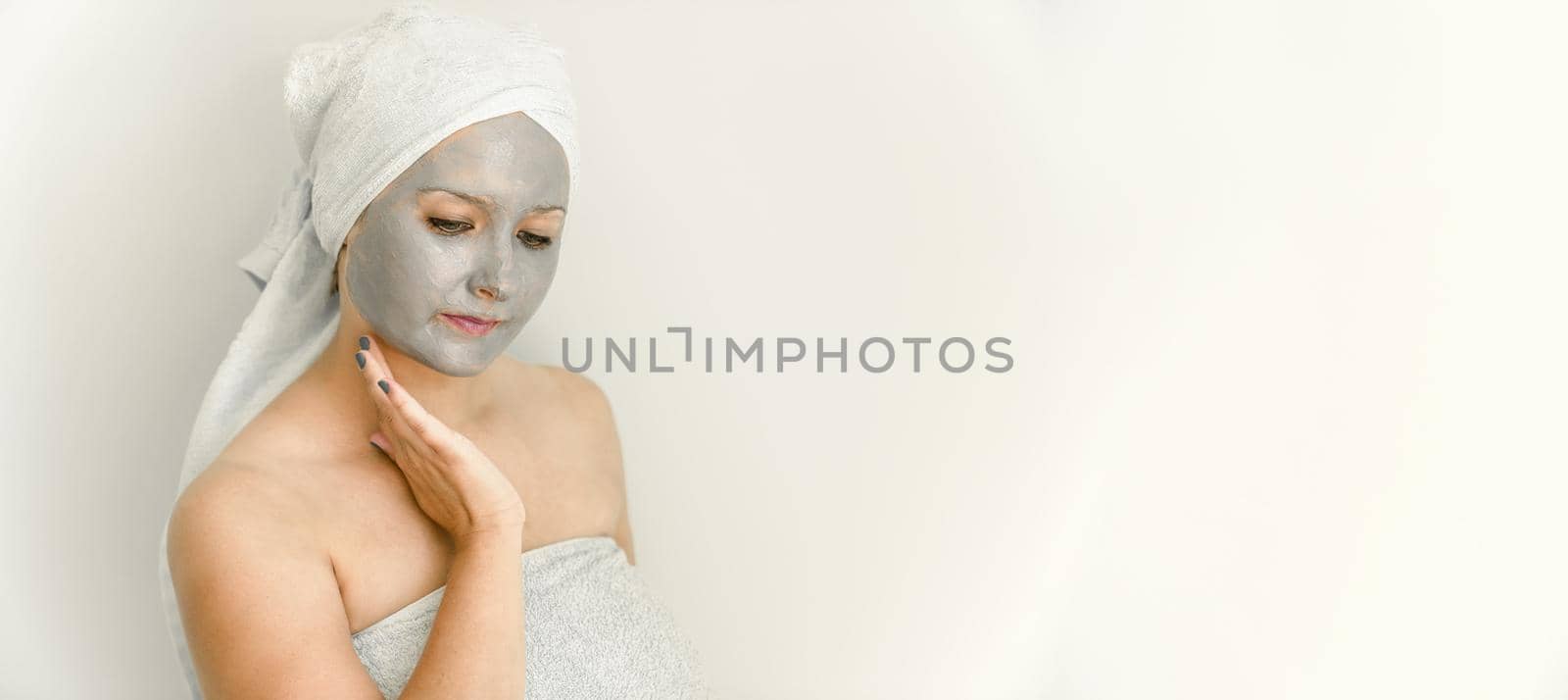 Young woman with clay cosmetic mask, wrapped in towel, touch face with hand, her gaze is directed downward. Facial skin care with cosmetic treatment. Copy space, banner