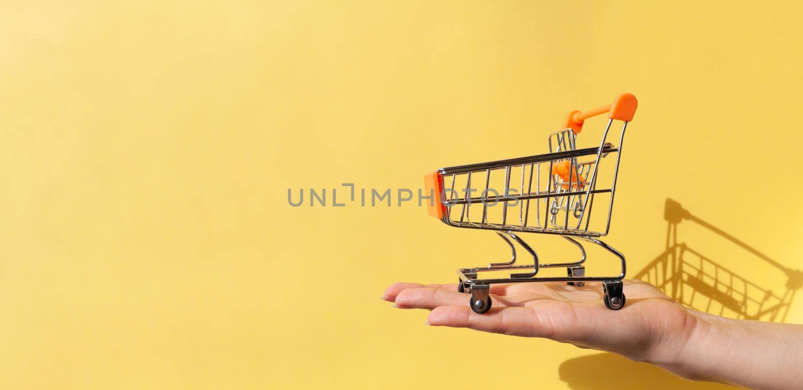 On a woman's hand is a small empty metal trolley from a supermarket. Banner. The object is on a yellow background. Horizontal photo. trading concept. Buyer. Shopping concept. Close-up. Copy space.