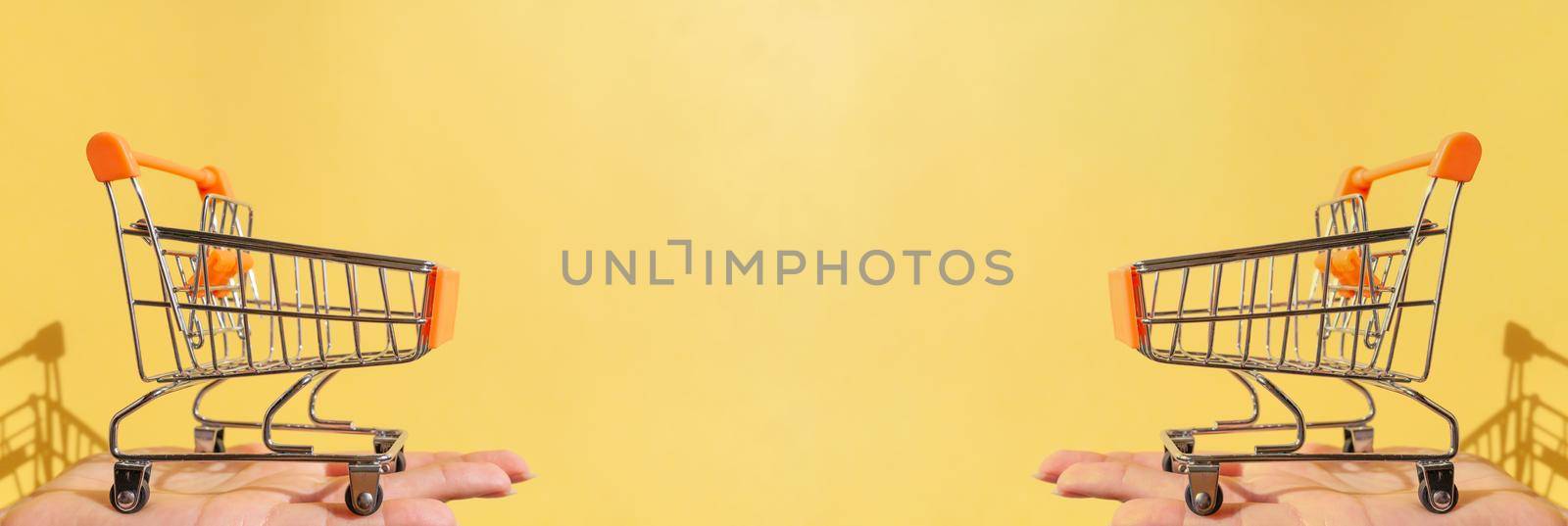 On a woman's hand is a small empty metal trolley from a supermarket. Banner. The object is on a yellow background. Horizontal photo. trading concept. Buyer. Shopping concept. Close-up. Copy space.
