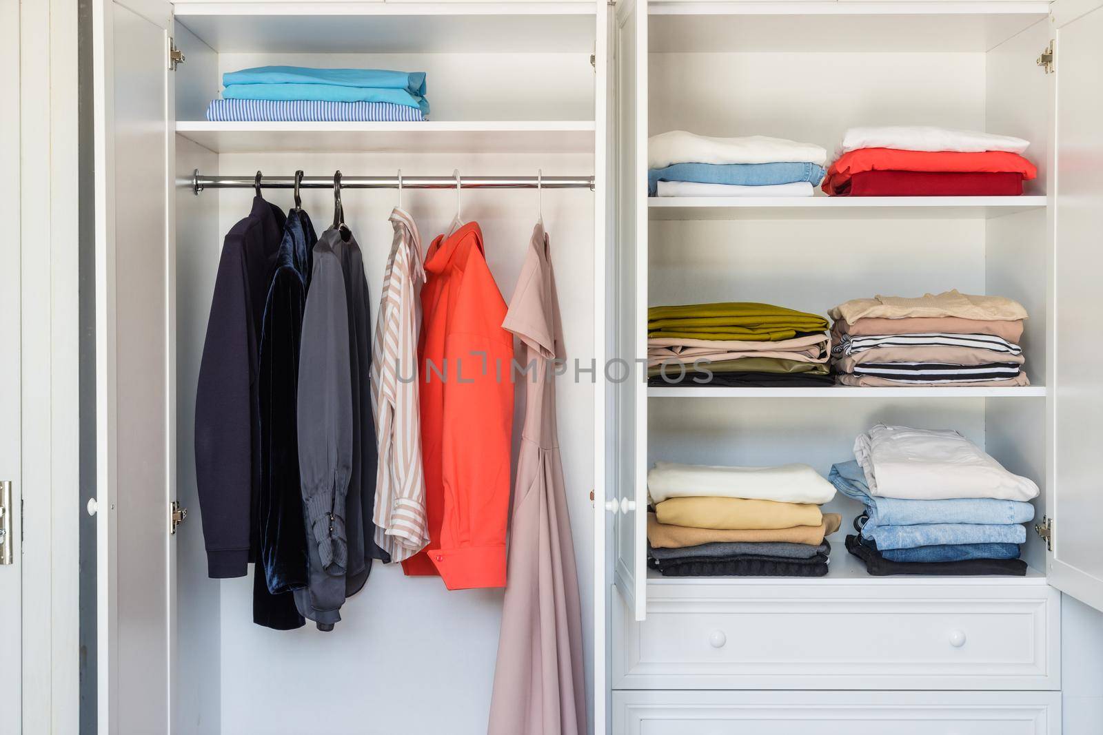 Close-up, open white closet in which blouses, dresses and jackets hang. Burdened minimalist approach to clothing selection. Working with a stylist to select a basic collection of things