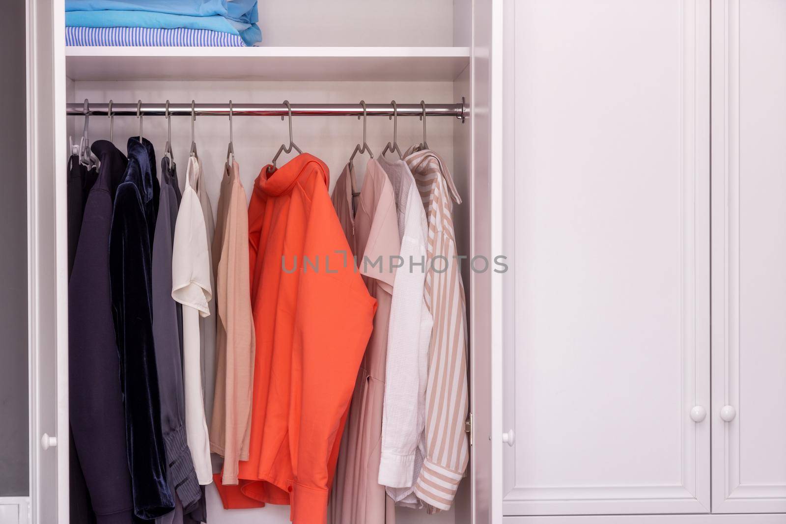 Close-up, open white closet in which blouses, dresses and jackets hang. Burdened minimalist approach to clothing selection. Working with a stylist to select a basic collection of things