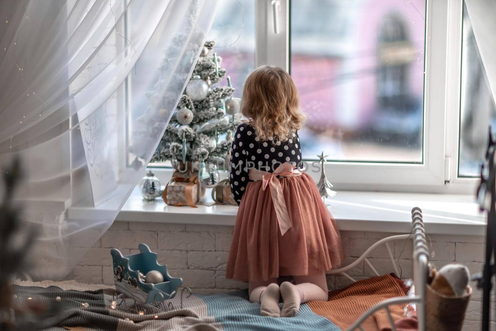 A little girl is sitting on the bed by the window and decorating a small tree with tiny Christmas toys. Happy healthy child celebrating a traditional family holiday. Adorable baby