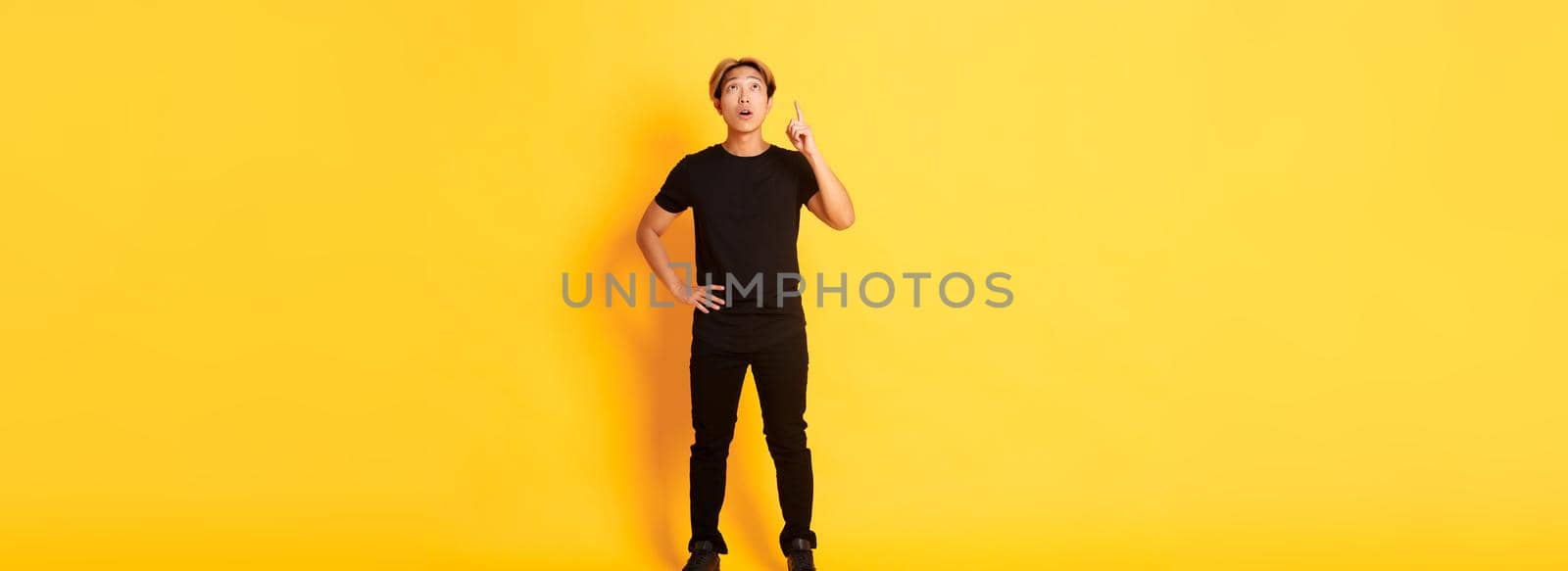 Full-length of thoughtful, intrigued attractive blond asian man raising finger, pointing up and looking amused, yellow background.