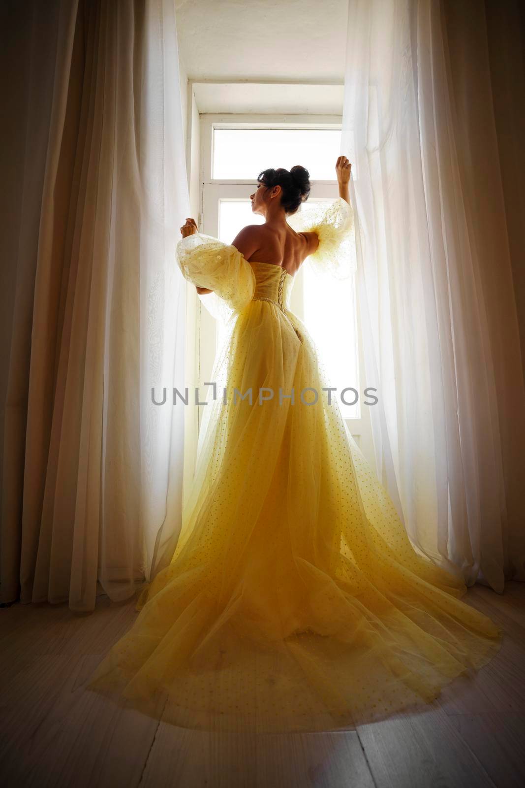 A woman's silhouette in a golden luxurious dress against the background of a window holds a curtain with her hands. Elegant lady in a yellow long silk dress with bare back, back view