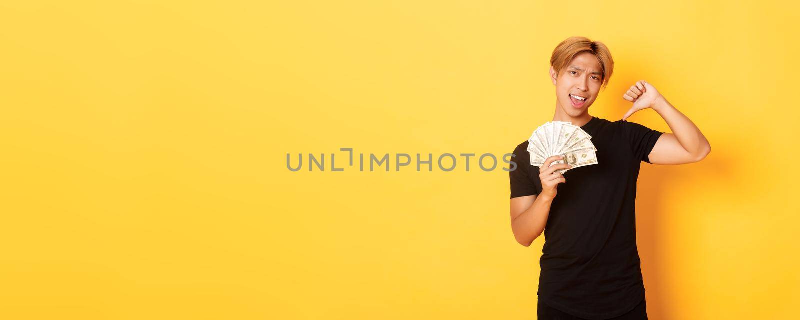 Sassy handsome asian guy pointing finger at cash and looking pleased. Korean man borrowed money, standing yellow background.