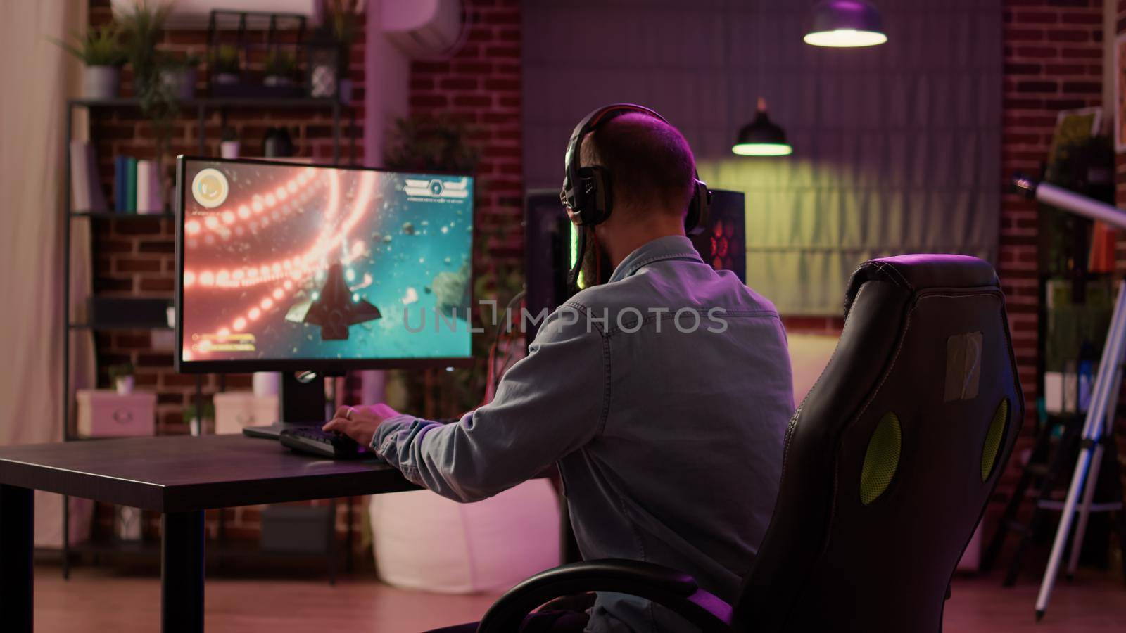 Over shoulder view of caucasian man using pc setup playing multiplayer online action game talking to team on headset. Gamer streaming fast paced space shooter while explaining gameplay to subscribers.