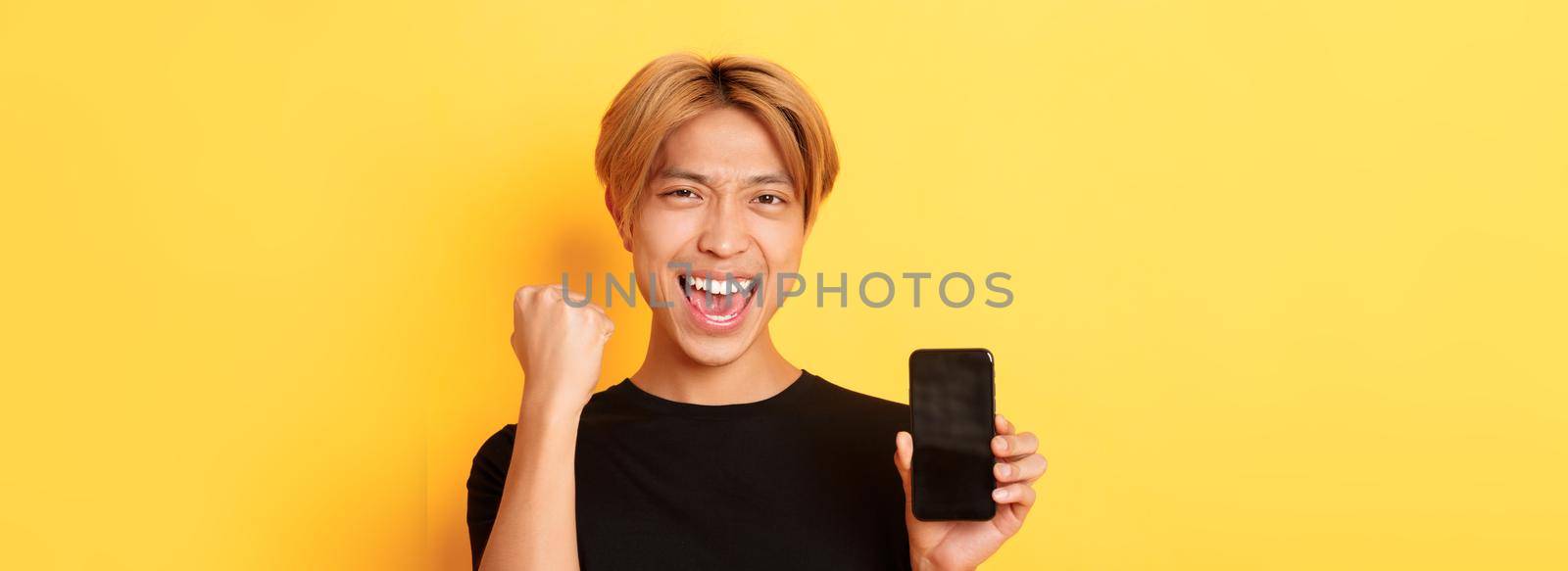 Close-up of happy rejoicing asian guy showing smartphone screen and saying yes, fist pump as triumphing, winning or achieve goal, yellow background.