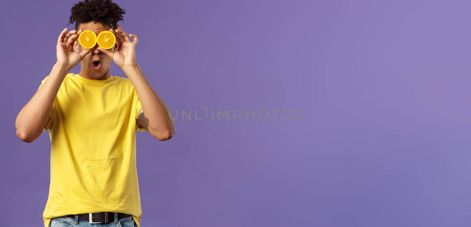 Holidays, vitamins and vacation concept. Portrait of funny, playful young guy fool around, playing with fruits, making eyes from pieces of orange, show wondered face expression, purple background by Benzoix