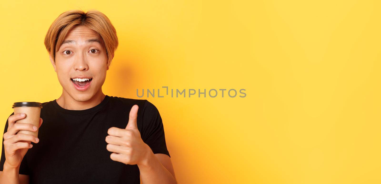 Close-up of surprised asian handsome guy recommend cafe, holding cup of coffee and showing thumbs-up in approval, smiling pleased over yellow background.