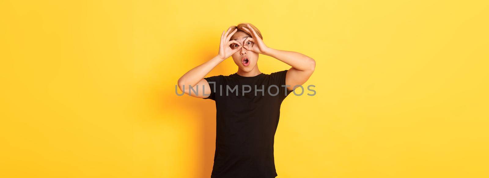 Portrait of funny and cute asian blond guy making fingers glasses and grimacing, standing yellow background.