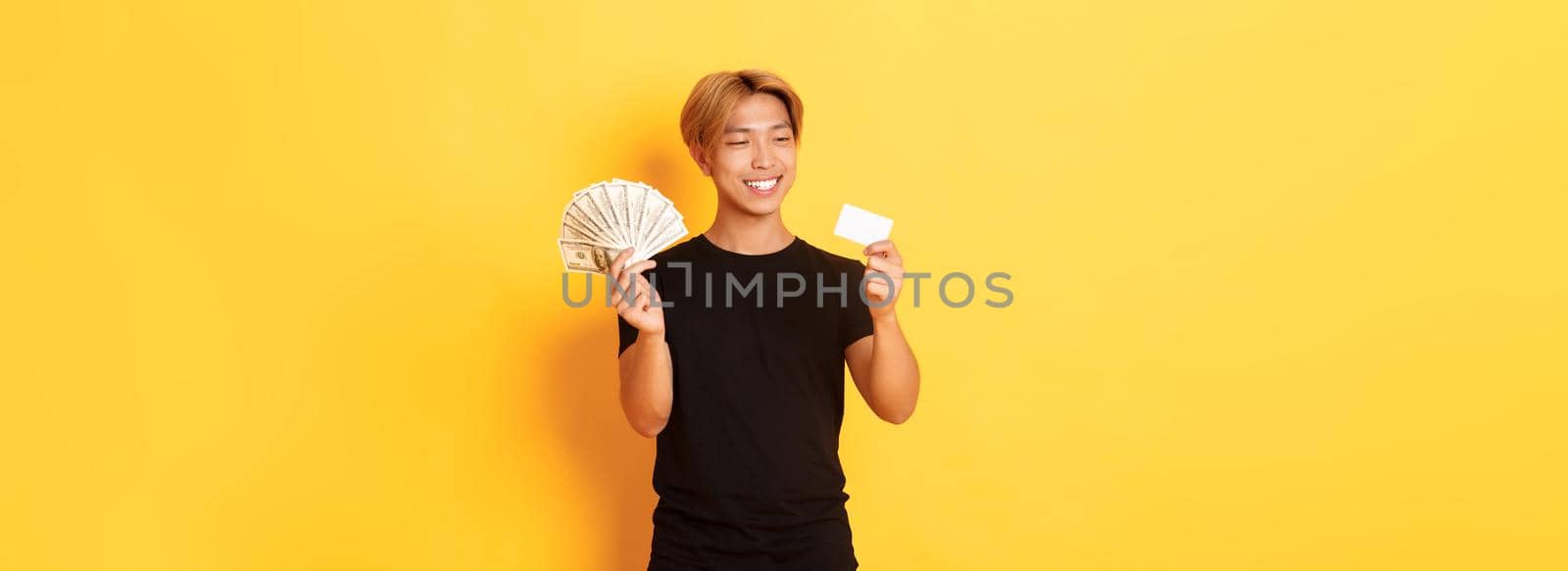 Portrait of satisfied smiling, handsome asian guy looking pleased at credit card while also holding money, standing yellow background by Benzoix