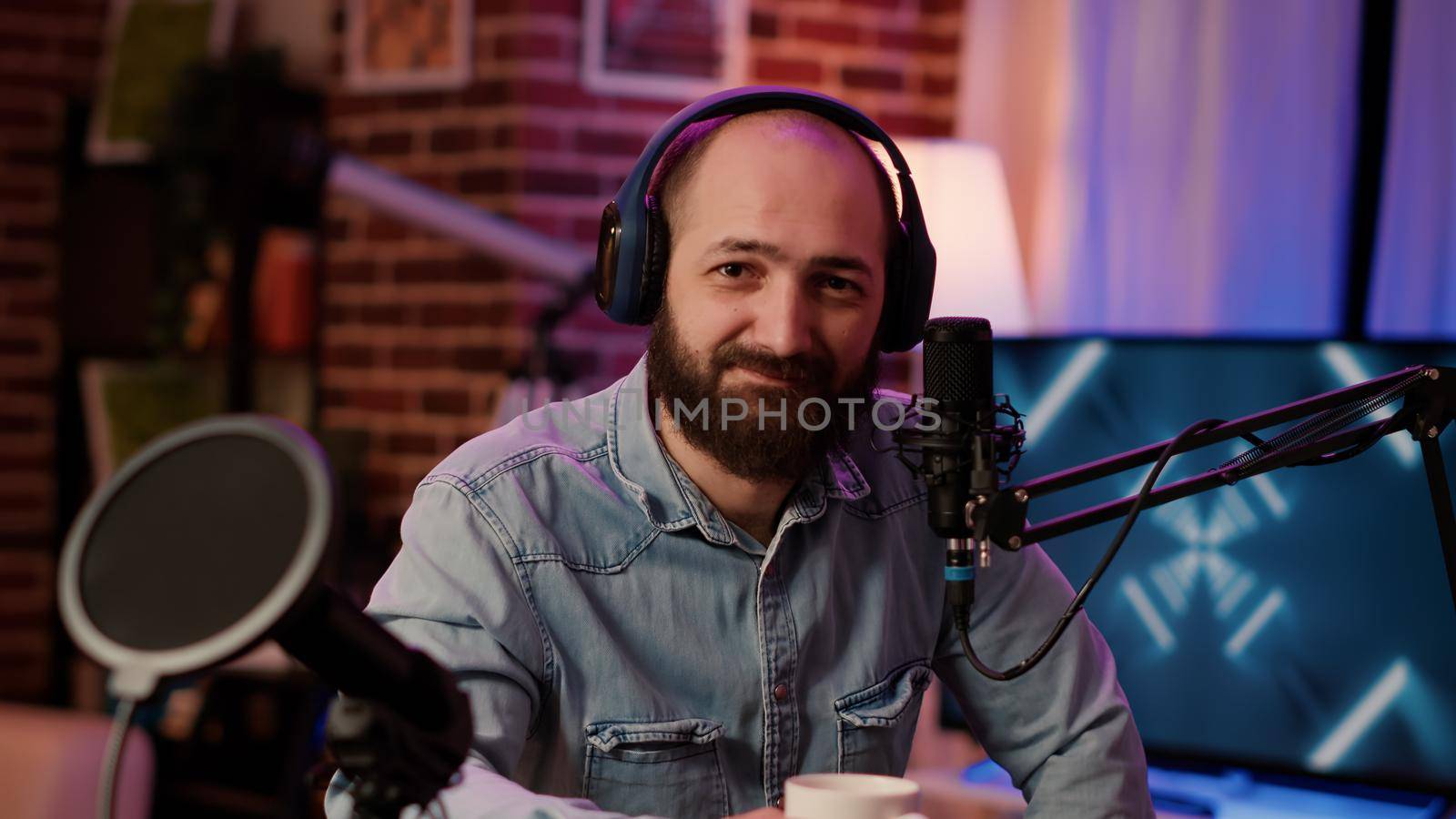 Closeup portrait of man recording online podcast with professional microphone smiling at camera sitting at desk. Online radio host with headphones streaming internet live show from home studio.