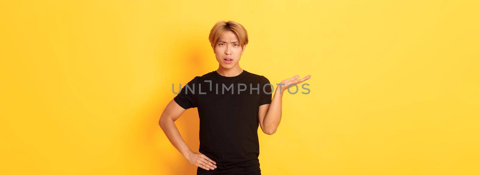 Frustrated and annoyed asian guy with blond hair, raising hand confused and looking at camera, yellow background.