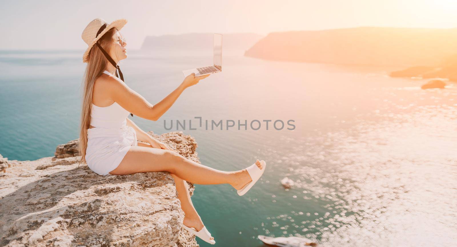 Successful business woman in yellow hat working on laptop by the sea. Pretty lady typing on computer at summer day outdoors. Freelance, travel and holidays concept.