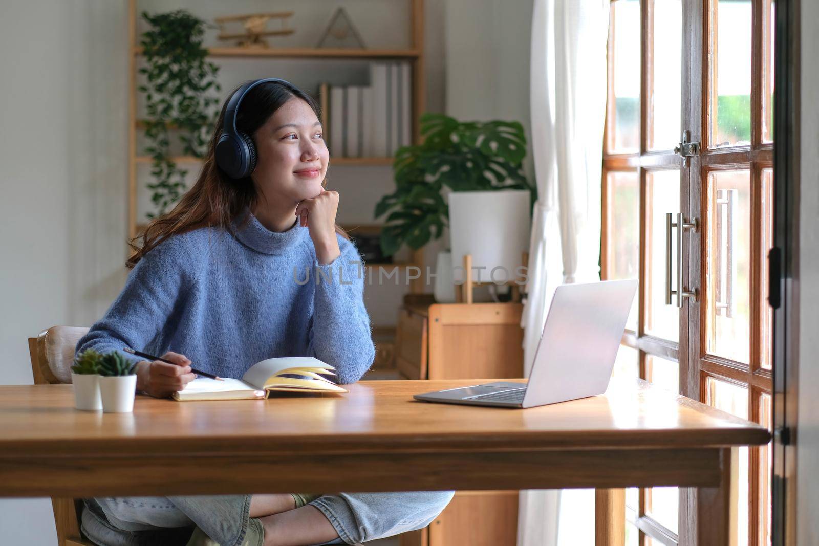 Smiling girl in headphones sitting at a desk looking outside and using a laptop to study online. Smart young women who are happy in headphones take courses on the web or practice using computers.