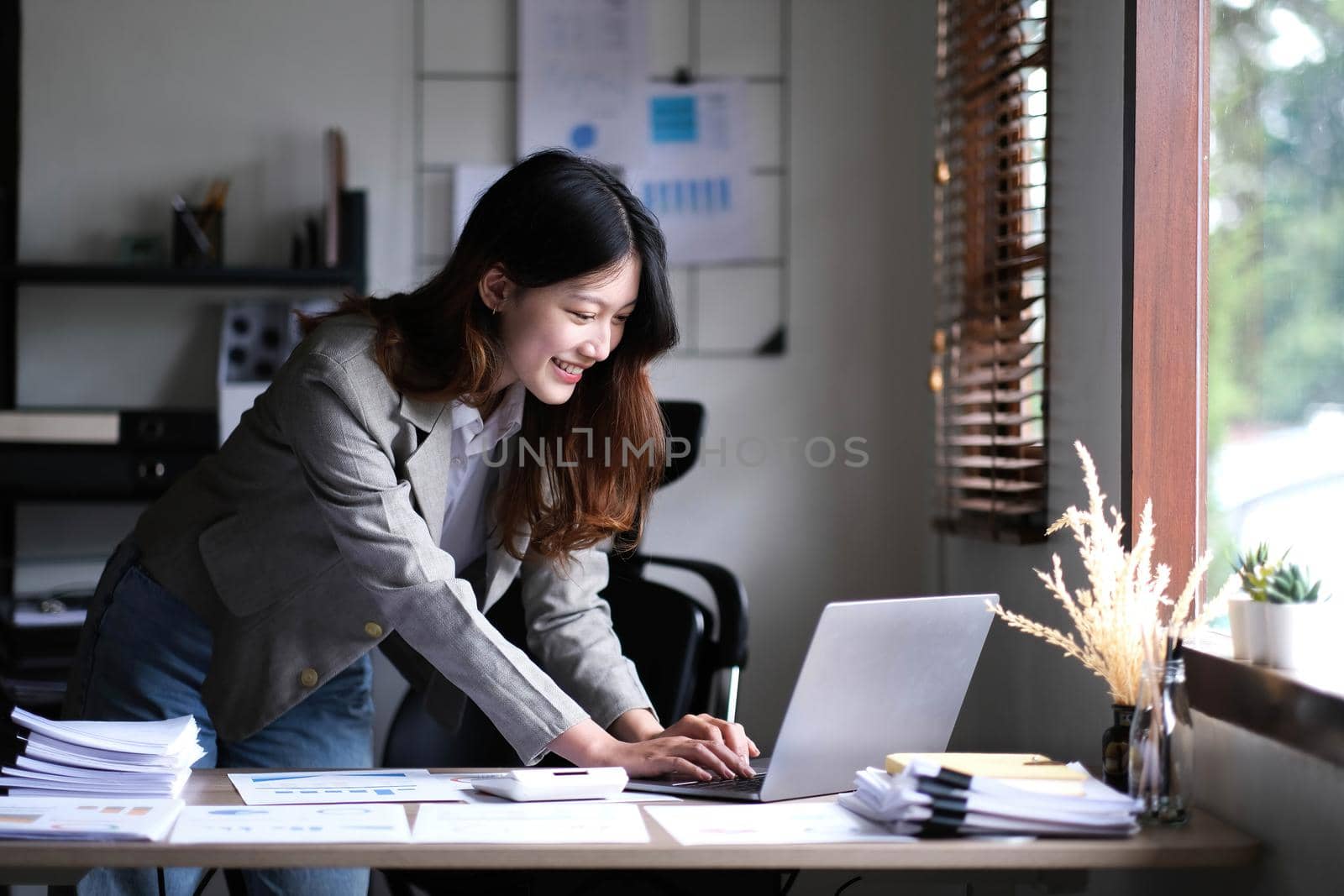 Happy young Asian businesswoman standing using calculator and laptop computer at office..