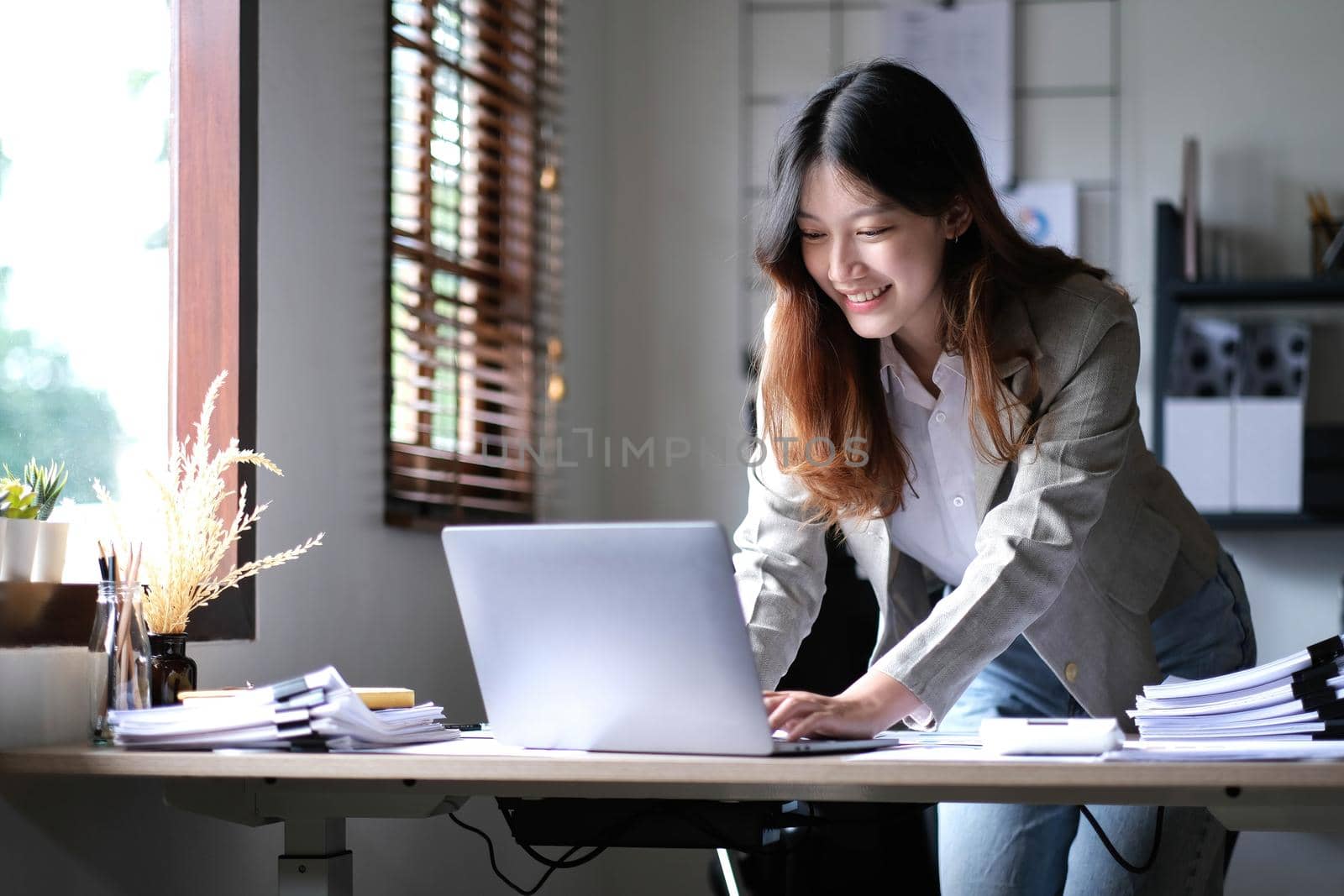 Beautiful Asian businesswoman standing and working on laptop with smiling face while working in office..