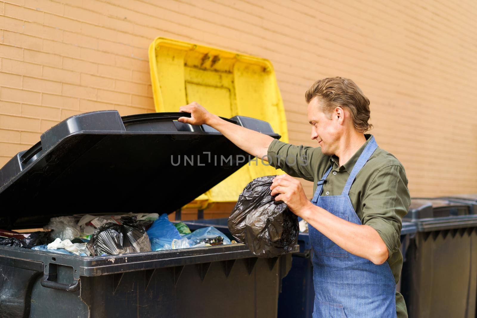 Caucasian handsome man employee open can full of garbage to throw plastic bag with trash wearing apron. Wrong unsorted garbage cans. Wrong way to throw your trash. Sorting garbage save the world.