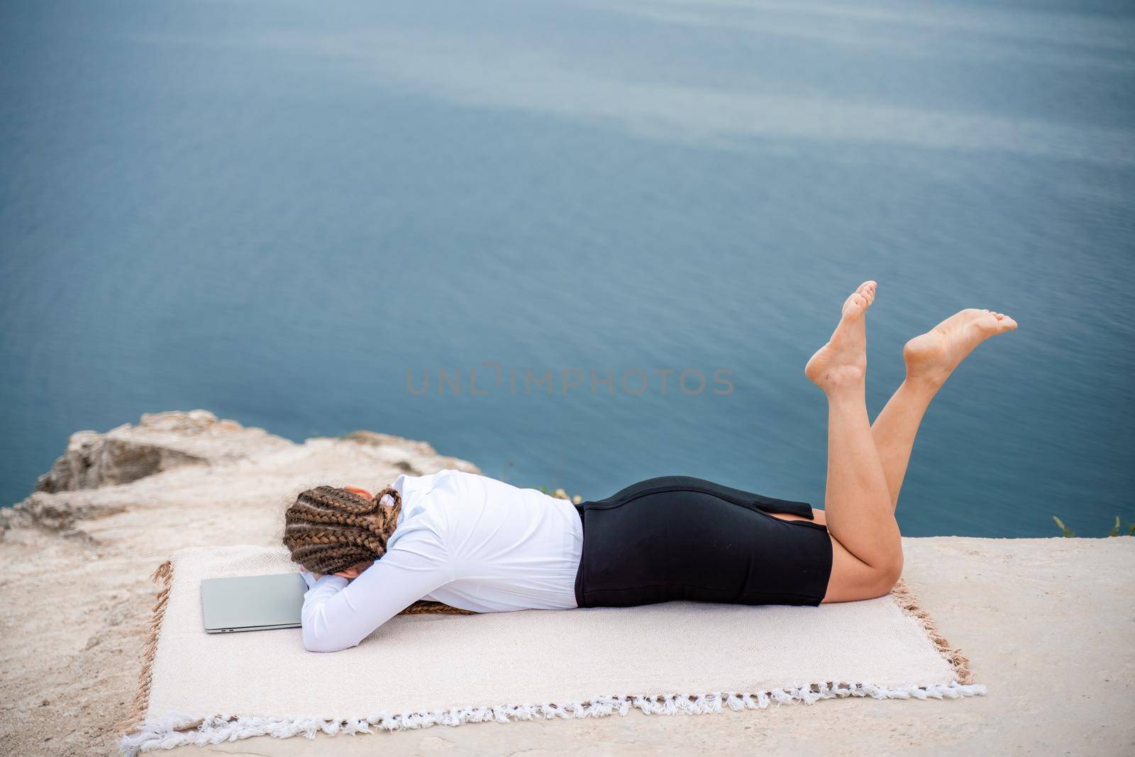 A woman is lying and typing on a laptop keyboard on a terrace with a beautiful sea view. Wearing a white blouse and black skirt. Freelance travel and vacation concept, digital nomad