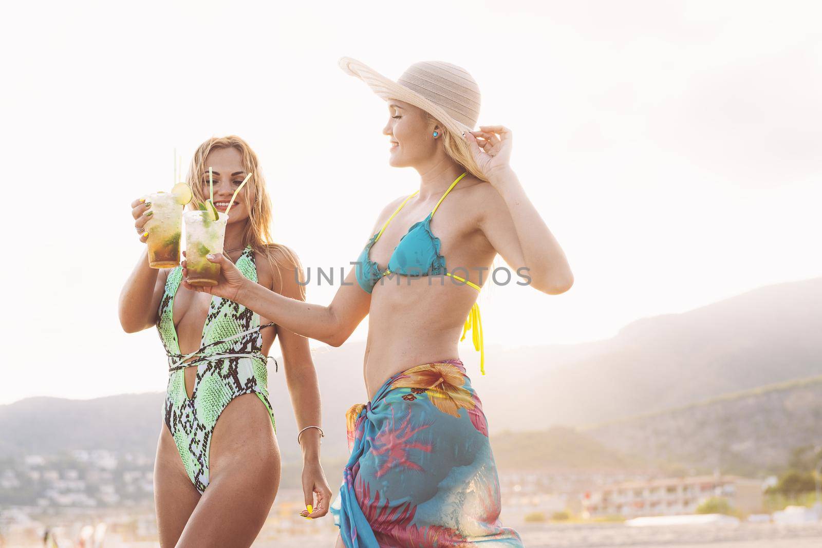 couple of two blonde girls toasting with mojitos on the beach at sunset, concept of friendship and relaxation in the summer