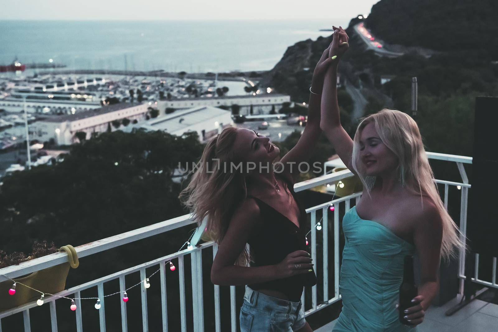 two beautiful young girls with beers dancing and celebrating at a private party on the outdoor terrace at the night, leisure happiness and friendship concept, vintage look with grain