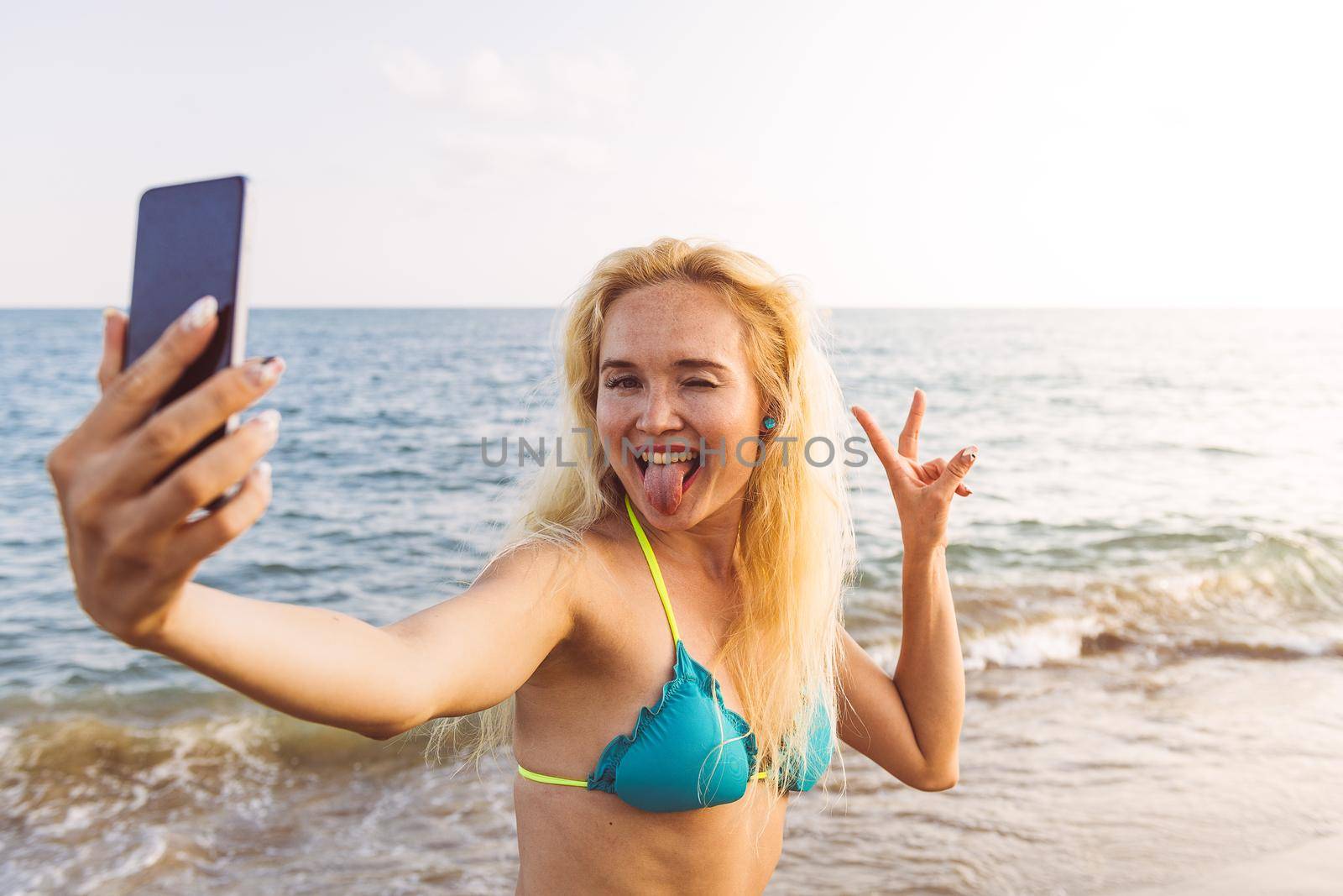 blonde woman taking a selfie photo with her mobile phone at beach at sunset on her summer vacation, communication and technology concept