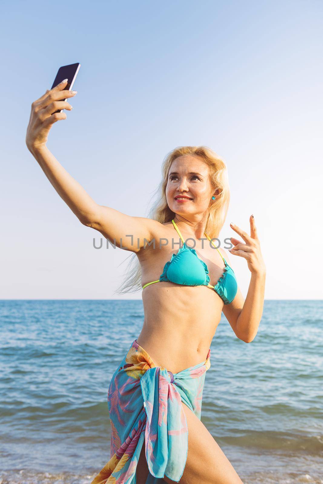 vertical photo of a blonde woman taking a selfie photo with her mobile phone at beach at sunset on her summer vacation, communication and technology concept