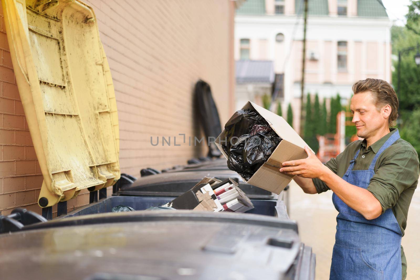 Throwing away unsorted trash employee man wearing apron put cardboard box with garbage plastic bag in it. Wrong unsorted garbage cans. Wrong way to throw your trash. Sorting garbage save the world.
