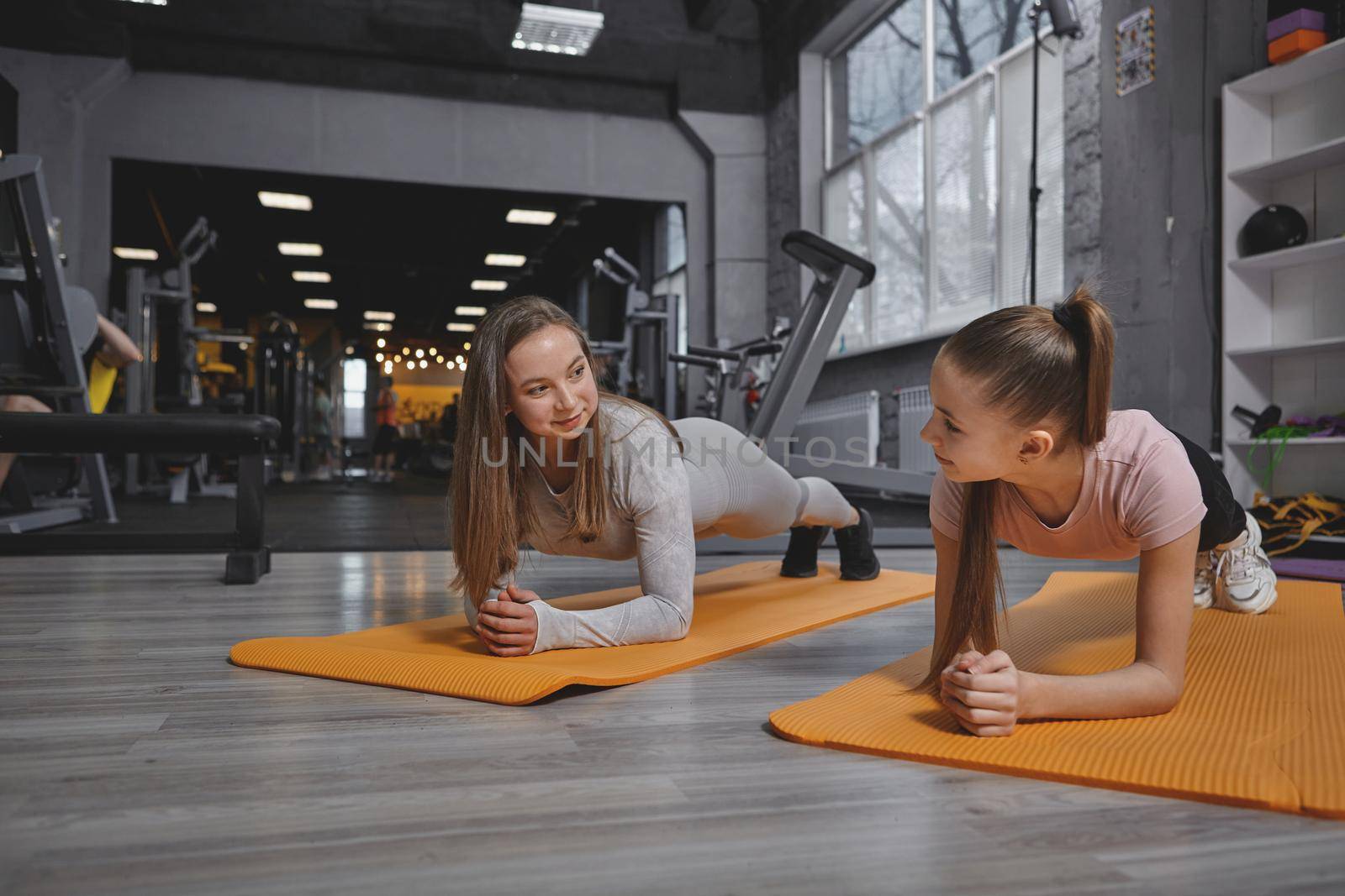 Lovely teenage girl smiling at her personal trainer while doing plank exercise together at the gym, copy space