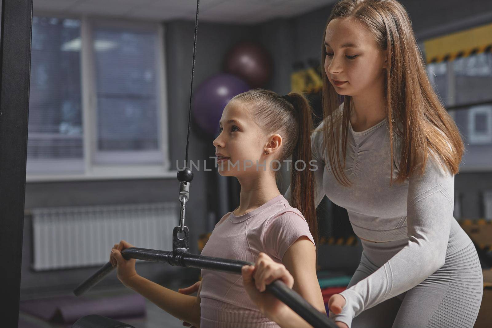 Focused teenage girl exercising on lat pulldown gym machine with her personal trainer