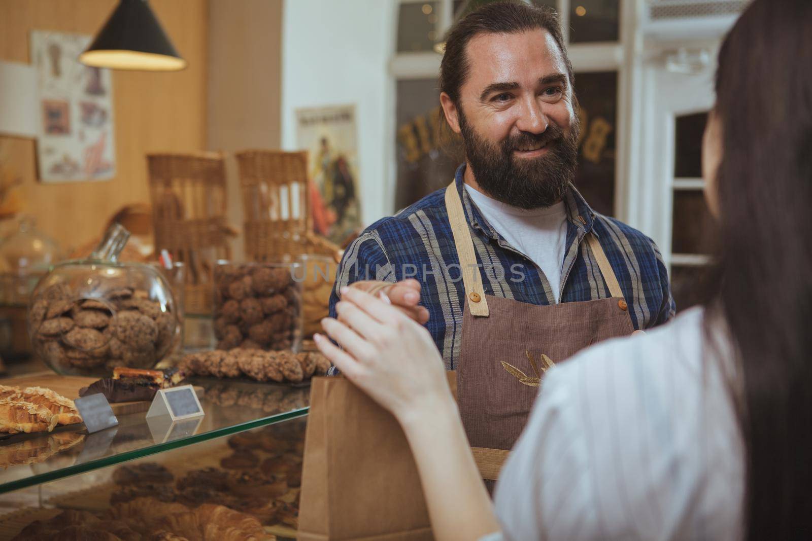 Handsome bearded mature baker wearing apron giving paper shopping bag to his customer, copy space. Unrecognizable woman shopping at bakery store, receiving her purchased pastry in a bag