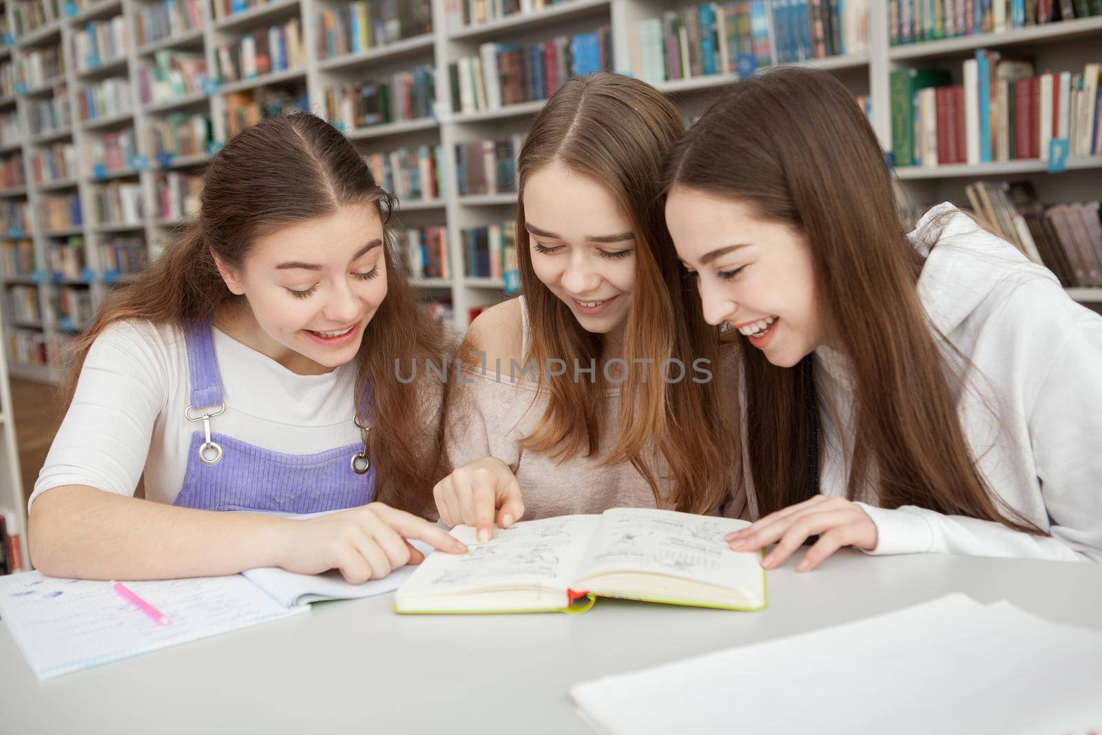 Cheerful teenage girls laughing while reading a book together at school library
