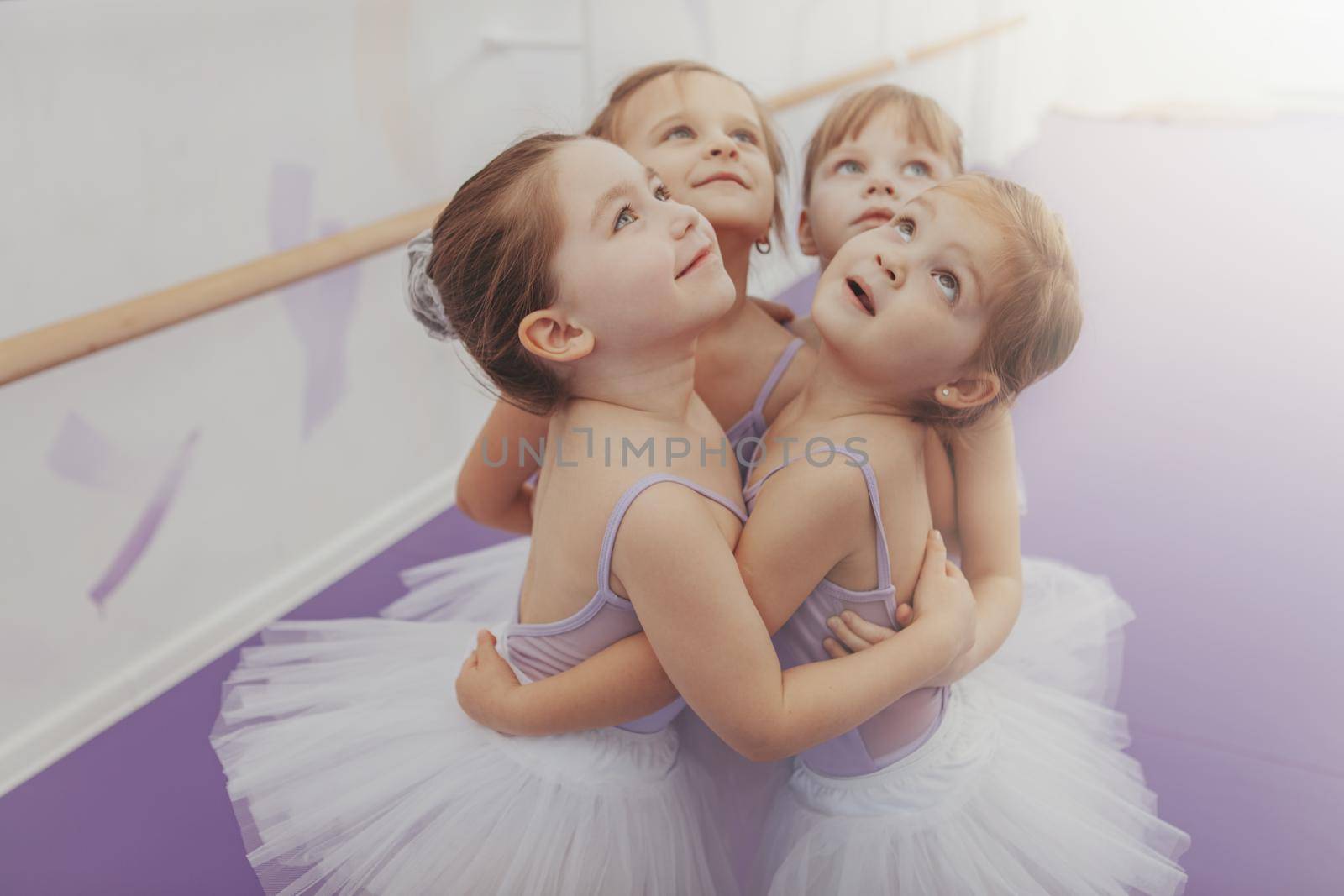 Adorable little girls in leotards and tutu skirts embracing, looking up joyfully. Group of cute young ballerinas having fun after ballet class, copy space. Friendship, childhood, sisterhood concept