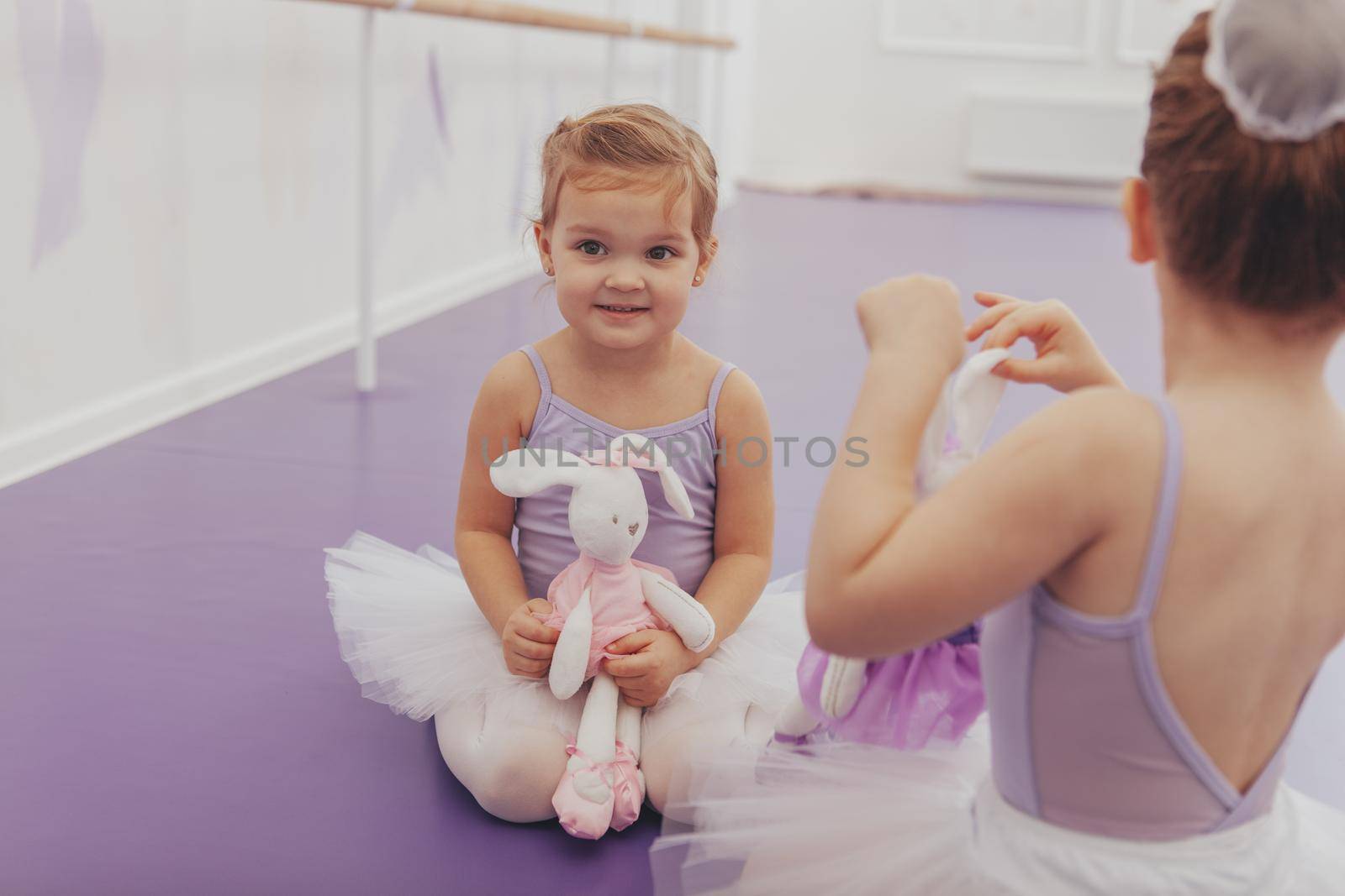 Cute happy little girl in leotard and tutu holding her toy, sitting with her best friend at ballet school, waiting for lesson. Little cute ballerinas relax before exercising at dance studio