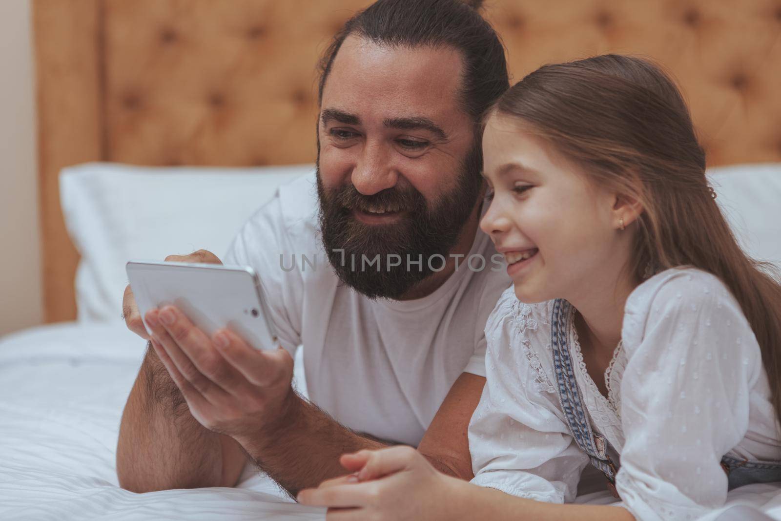 Close up of a handsome bearded mature man enjoying using smart phone with his adorable little daughter. Single father teaching his child how to use internet safely. Online, child safety concept