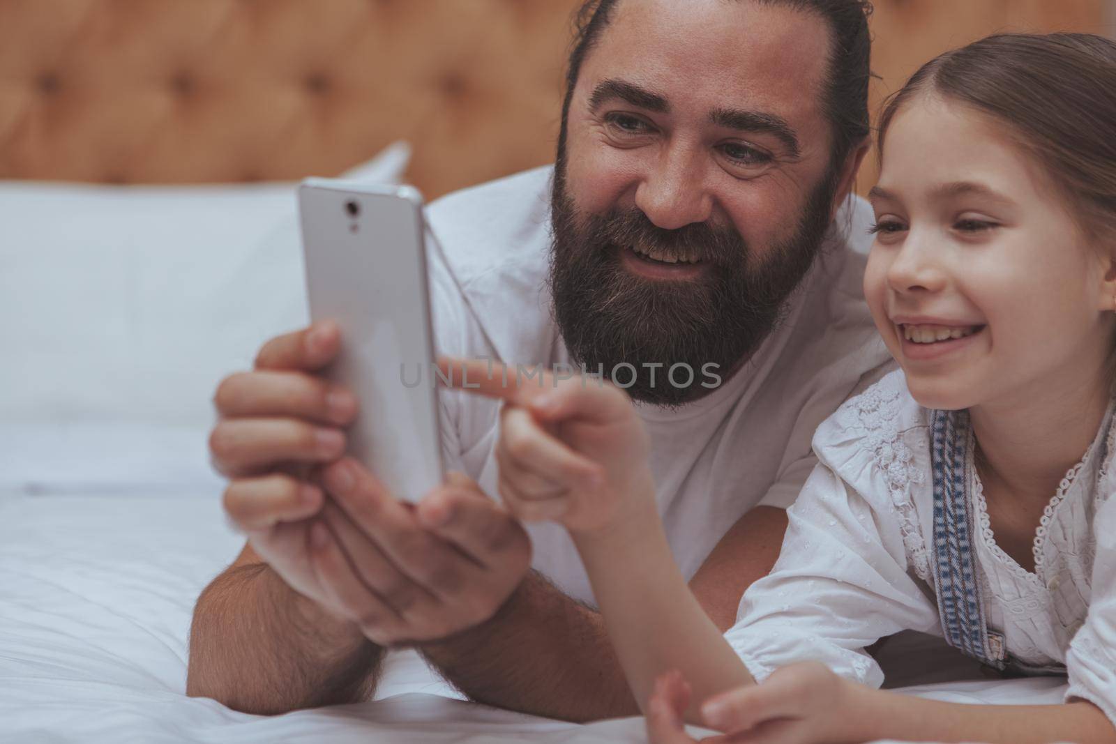 Cropped close up of a beautiful cheerful little girl browsing online, using smart phone with her loving father. Handsome bearded man smiling, enjoying spending weekend at home with his daughter, browsing online