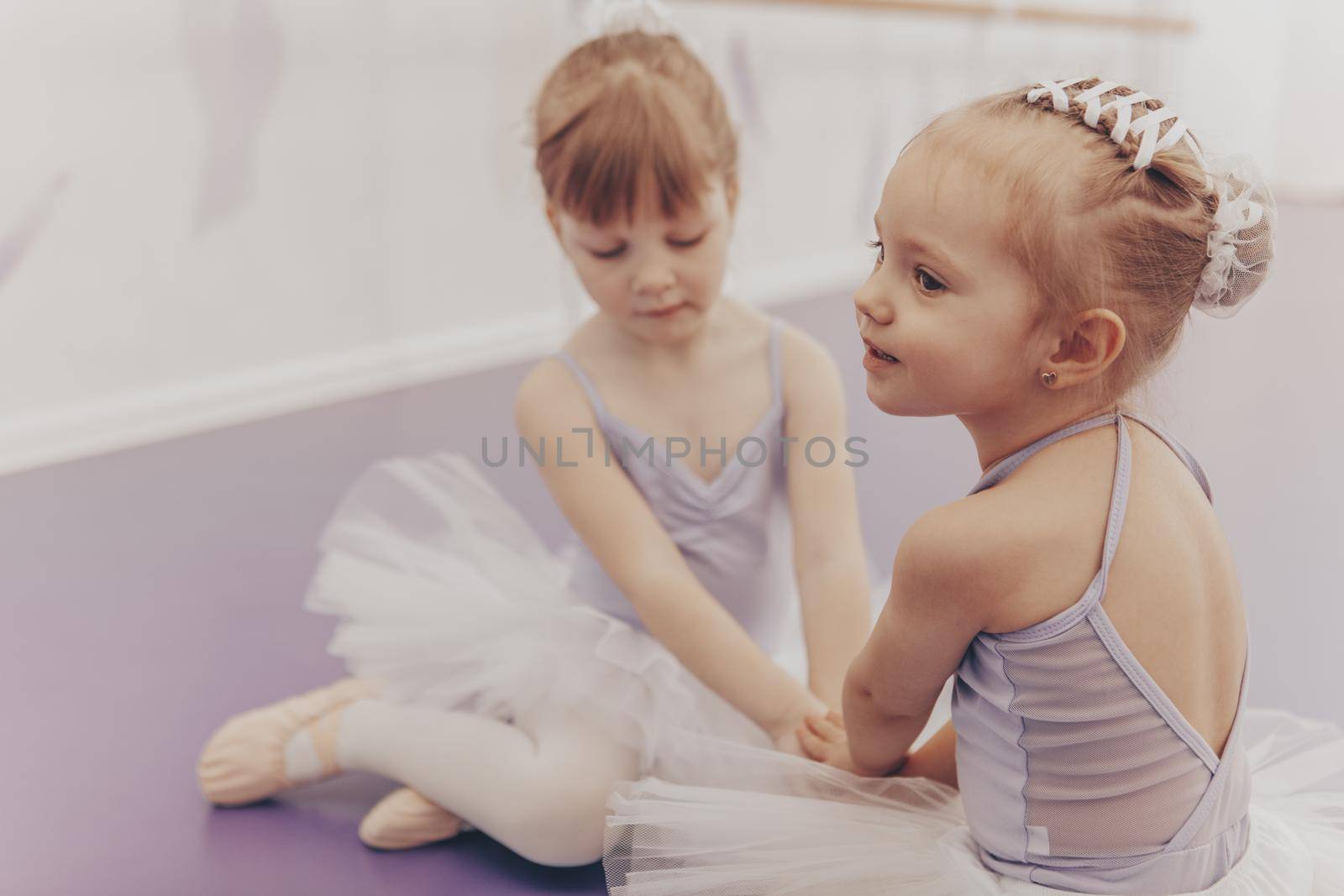 Lovely little girl sitting on the floor with her best friend at ballet school. Cute little ballerinas in leotards and tutu skirts resting after dancing, copy space. Friendship, innocence concept