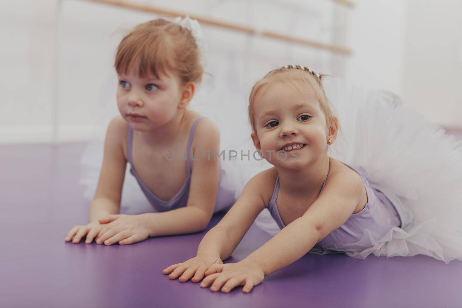 Beautiful happy little ballerina smiling to the camera lying on the floor with her best friend at ballet school. Two lovely little girls in ballet dancing outfits relax after exercising